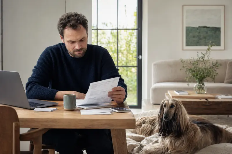 Ein Besitzer sitzt entspannt am Tisch mit einer Tierarztrechnung, während ein ruhiger Afghanischer Windhund auf einer Decke liegt.