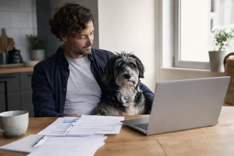 Ein Aidi Hund sitzt ruhig neben seinem Besitzer am Küchentisch, auf dem Versicherungsunterlagen und ein Laptop liegen.