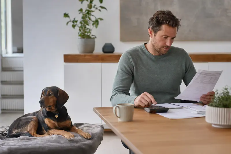 Ein Besitzer mit einer Tierarztrechnung sitzt entspannt am Tisch, während sein Alpenländische-Erzgebirgler Dachsbracke neben ihm auf einer Decke liegt.