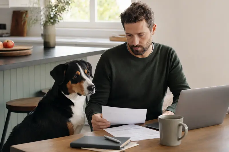 Ein Appenzeller Sennenhund sitzt entspannt neben seinem Besitzer am Küchentisch, auf dem sich Versicherungsunterlagen und ein Laptop befinden.