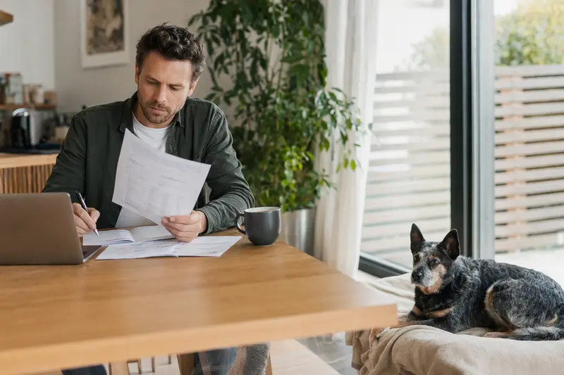 Besitzer am Tisch mit Tierarztrechnung, während ein entspannter Australian Cattle Dog auf einer Decke liegt.