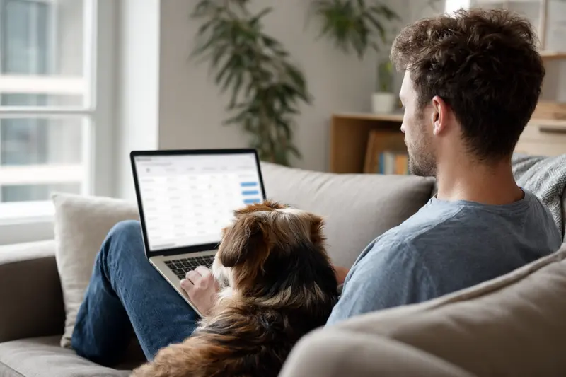 Besitzer sitzt mit Laptop auf dem Sofa, neben ihm ein Bakarwal Hund in einer hellen Wohnung.