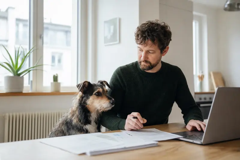 Ein Barb Hund sitzt neben seinem Besitzer am Küchentisch, während Versicherungsunterlagen und ein Laptop auf dem Tisch liegen.