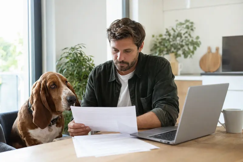 Ein Basset Hound sitzt entspannt neben seinem Besitzer am Küchentisch, umgeben von Versicherungsunterlagen und einem Laptop.