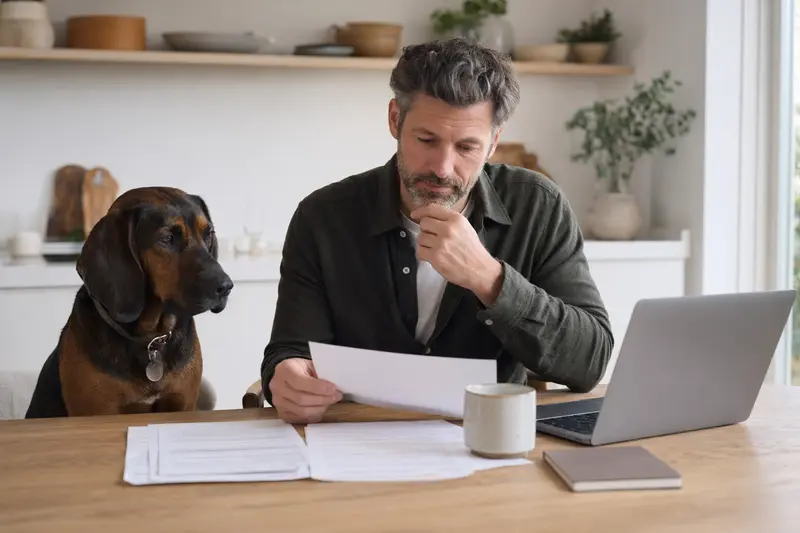 Ein Bayerischer Gebirgsschweißhund sitzt neben seinem Besitzer am Küchentisch, auf dem Versicherungsunterlagen und ein Laptop liegen.