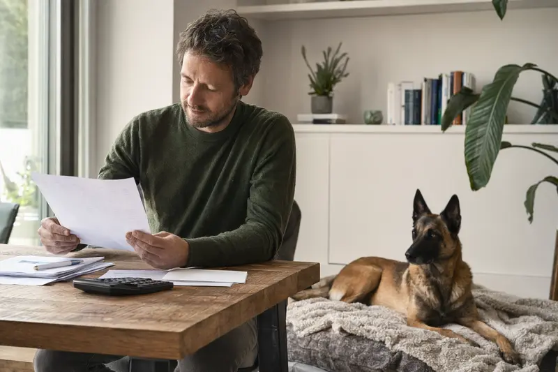 Ein Besitzer sitzt mit einer Tierarztrechnung am Tisch, während ein entspannter Belgischer Schäferhund neben ihm auf einer Decke liegt – ein ruhiges Bild zum Thema Hundekrankenversicherung und Tierarztkosten.