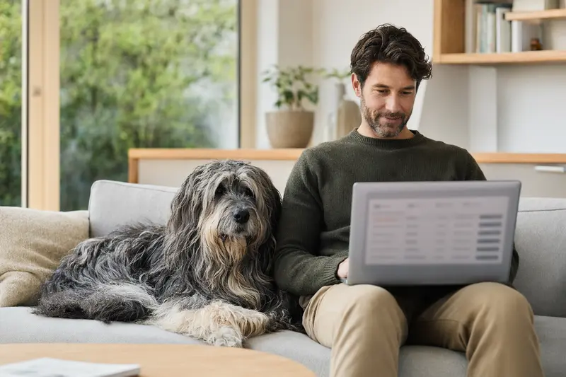 Ein Hundebesitzer sitzt mit einem Laptop auf dem Sofa, während ein Bergamasker Hirtenhund neben ihm liegt.