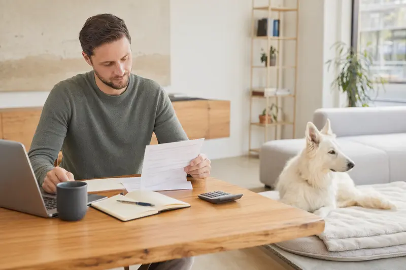 Ein Hundehalter sitzt mit einer Tierarztrechnung am Tisch, während ein entspannter Berger Blanc Suisse auf einer Decke daneben liegt – ein ruhiges Motiv zum Thema Tierarztkosten und Hundeversicherung.