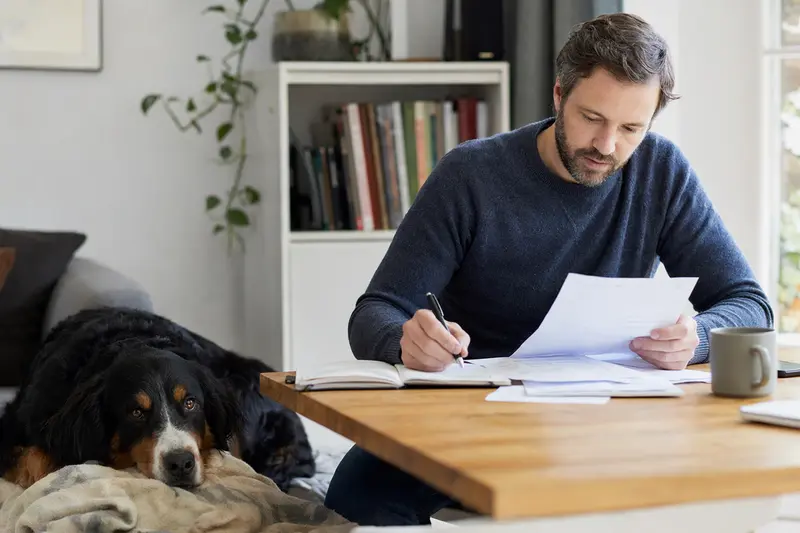 Ein Hundehalter sitzt mit einer Tierarztrechnung am Tisch, während ein entspannter Berner Laufhund auf einer Decke liegt – ein ruhiges Motiv zum Thema Hundekrankenversicherung und Tierarztkosten.