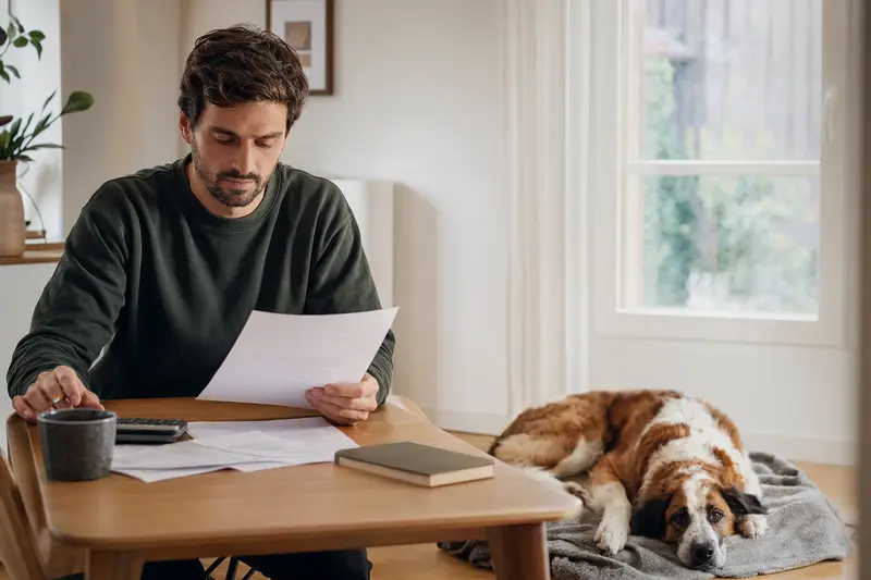 Ein Besitzer sitzt mit einer Tierarztrechnung am Tisch, während ein entspannter Bernhardiner auf einer Decke liegt – ein realistisches Bild zum Thema Tierarztkosten und Hund versichern.