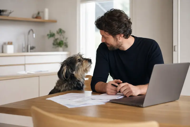 Ein Billy Hund sitzt friedlich neben seinem Besitzer am Küchentisch, während Versicherungsunterlagen und ein Laptop auf dem Tisch liegen.