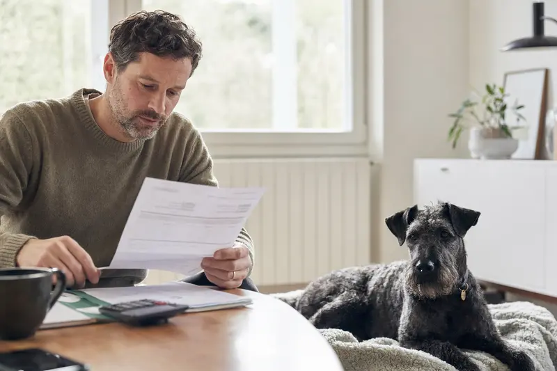 Ein Hundebesitzer sitzt mit einer Tierarztrechnung am Tisch, während ein Blauer Irischer Terrier entspannt auf einer Decke neben ihm liegt.
