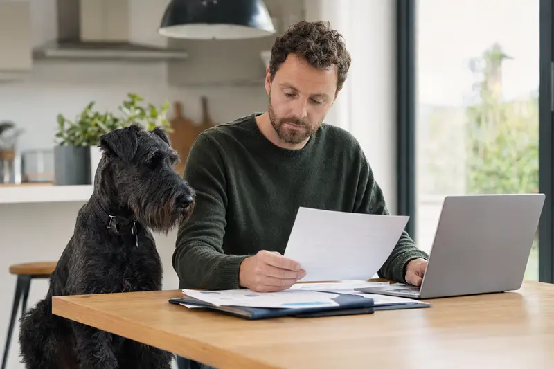 Ein blauer Irischer Terrier sitzt ruhig neben seinem Besitzer am Küchentisch, während Versicherungsunterlagen und ein Laptop mit Vergleichsseite auf dem Tisch liegen.
