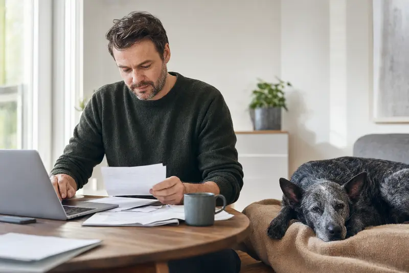 Ein Hundehalter sitzt mit einer Tierarztrechnung am Tisch, während ein entspannter Blue Lacy Hund ruhig auf einer Decke liegt.