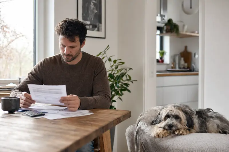 Besitzer sitzt mit Tierarztrechnung am Tisch, während ein entspannter Bobtail auf einer Decke liegt – ein ruhiges Motiv zum Thema Hundekrankenversicherung und Tierarztkosten.