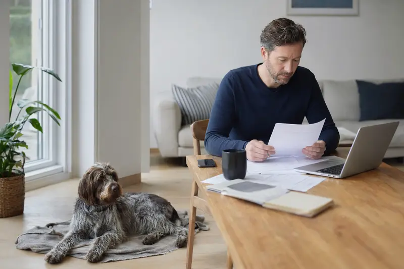 Ein Besitzer sitzt mit einer Tierarztrechnung am Tisch, während ein entspannter Böhmischer Rauhbart-Hund auf einer Decke liegt, passend zum Thema Hundekrankenversicherung.