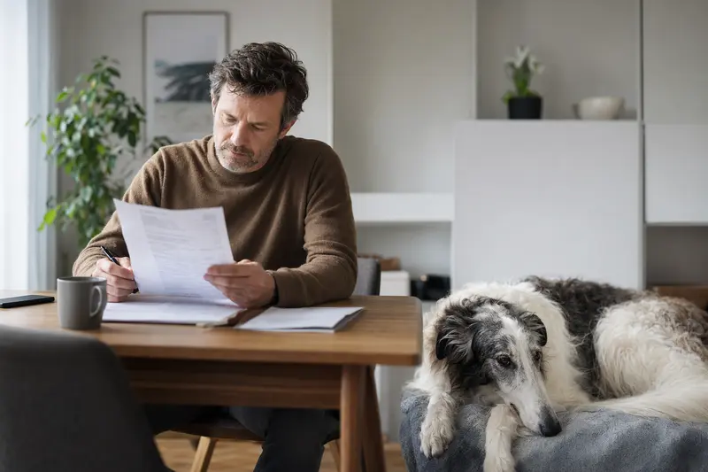 Ein Hundehalter sitzt ruhig am Tisch mit einer Tierarztrechnung, während ein entspannter Borzoi auf einer Decke daneben liegt.