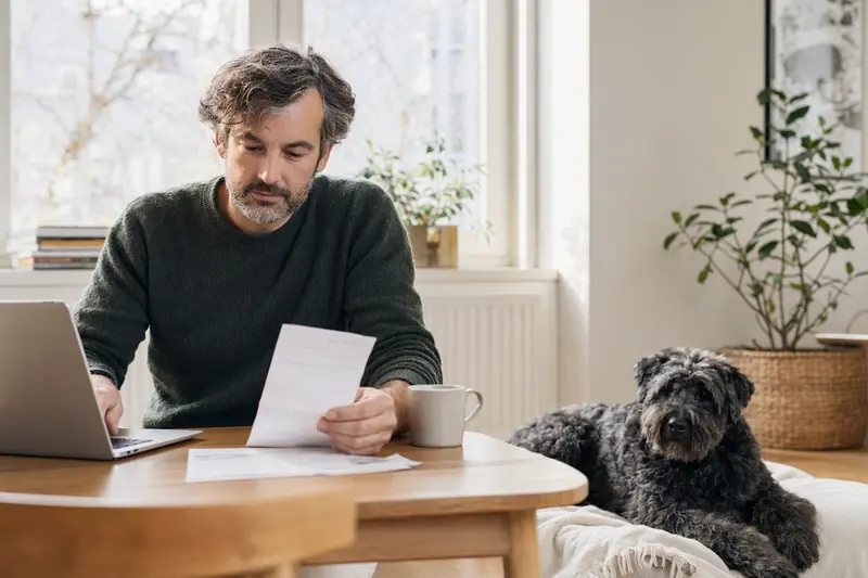 Ein Hundebesitzer sitzt mit einer Tierarztrechnung am Tisch, während ein entspannter Bouvier des Flandres auf einer Decke daneben liegt.