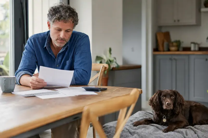 Ein Hundehalter sitzt mit einer Tierarztrechnung am Tisch, während ein entspannter Boykin Spaniel auf einer Decke liegt und die Situation im Zusammenhang mit der Hundeversicherung ruhig wirkt.