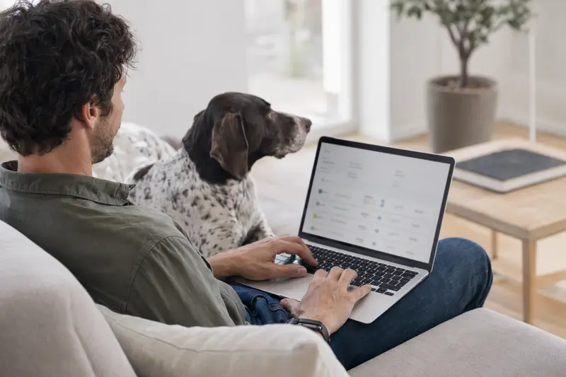 Ein Hundehalter sitzt mit einem Laptop auf dem Sofa, während sein Braque Francais Typ Pyrenees neben ihm liegt, passend zur Auswahl einer Tierkrankenversicherung.