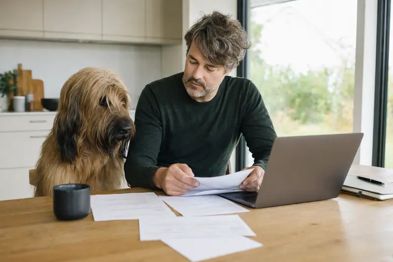 Ein Briard Hund sitzt neben seinem Besitzer am Küchentisch, wo Versicherungsunterlagen und ein Laptop liegen.