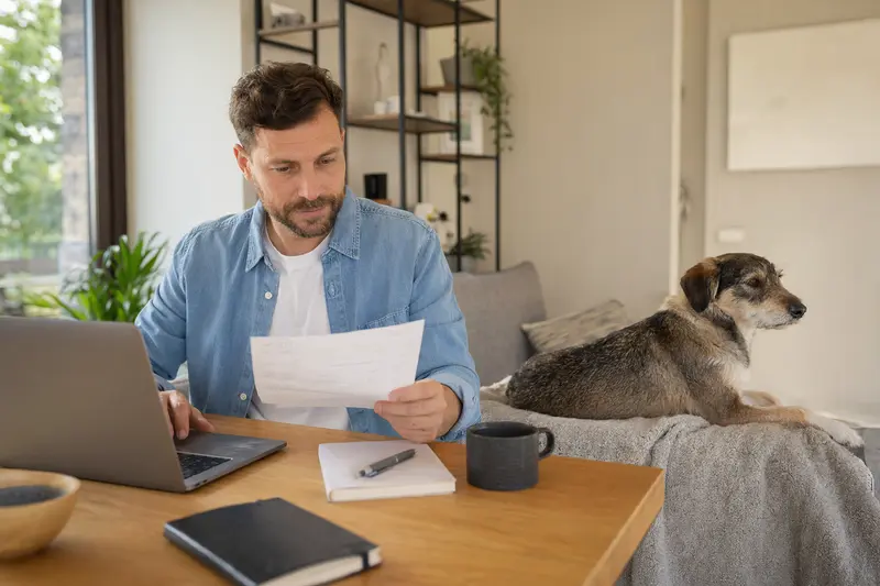 Ein Hundebesitzer sitzt mit einer Tierarztrechnung am Tisch, während ein entspannter Ca Eivissec Hund ruhig auf einer Decke neben ihm liegt.