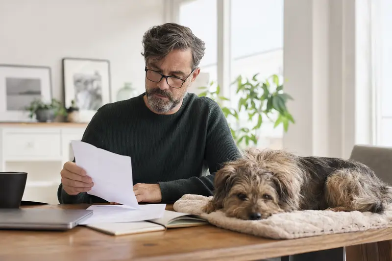 Ein Hundebesitzer sitzt mit einer Tierarztrechnung am Tisch, während ein entspannter Cairn Terrier auf einer Decke daneben liegt – ein Bild zum Thema Hundekrankenversicherung und Tierarztkosten.
