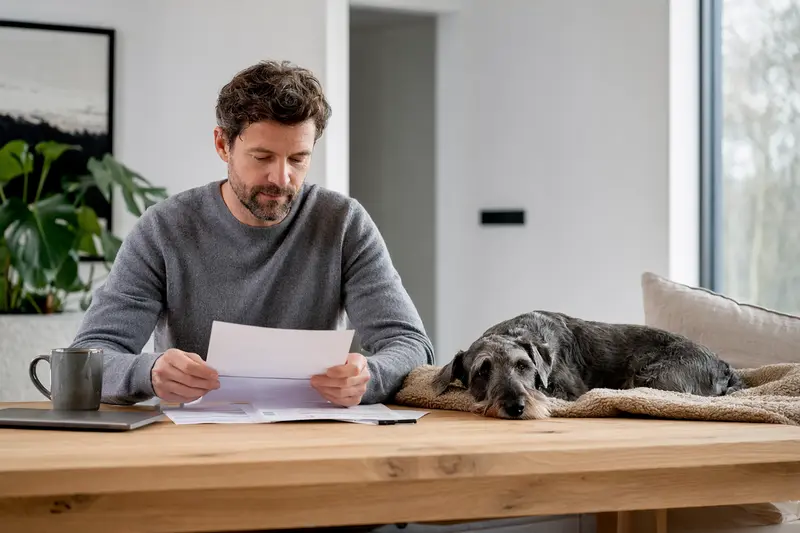Ein Cesky Terrier liegt ruhig auf einer Decke neben einem Besitzer, der am Tisch eine Tierarztrechnung prüft – ein realistisches Bild zum Thema Hundeversicherung.
