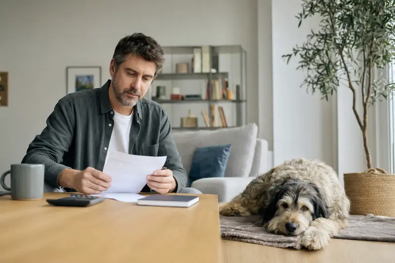 Ein Hundebesitzer sitzt mit einer Tierarztrechnung am Tisch, während ein Ciobanesc Romanesc Mioritic entspannt auf einer Decke liegt – ein ruhiges Motiv zum Thema Hundekrankenversicherung und Tierarztkosten.