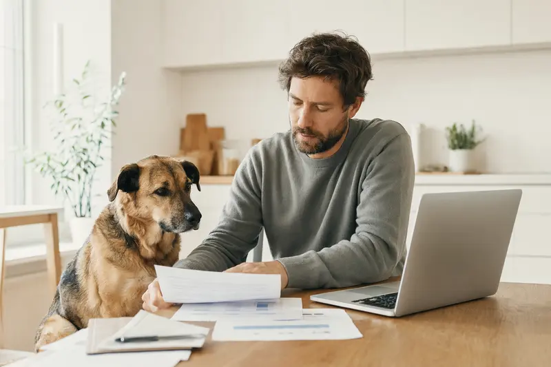 Ein Coban Hirtenhund sitzt ruhig neben seinem Besitzer am Küchentisch mit Versicherungsunterlagen und einem Laptop.