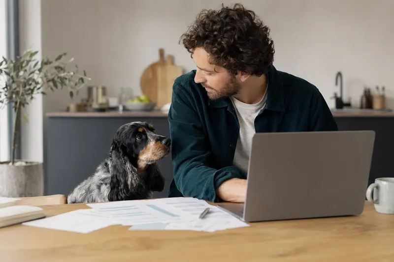 Cocker Spaniel sitzt ruhig neben seinem Besitzer am Küchentisch mit Versicherungsunterlagen und Laptop.