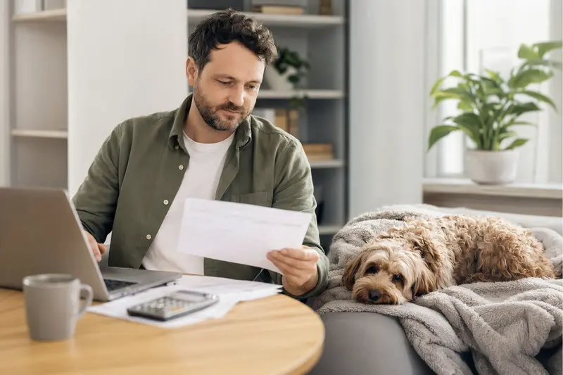 Ein Hundebesitzer sitzt mit einer Tierarztrechnung am Tisch, während sein entspannter Cockerpoo auf einer Decke liegt – ein ruhiges Motiv zum Thema Tierarztkosten und Hundekrankenversicherung.