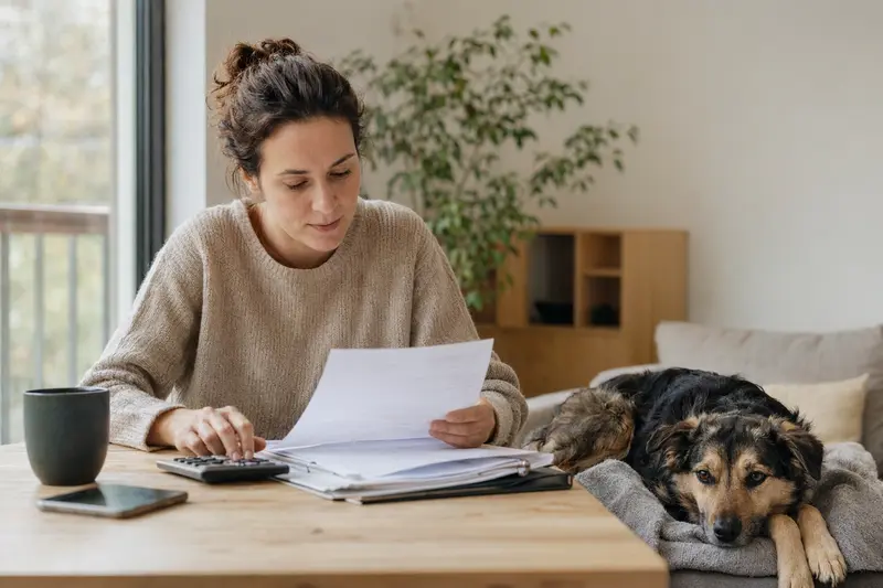 Ein Hundehalter sitzt mit einer Tierarztrechnung am Tisch, während ein entspannter Combai Hund auf einer Decke daneben liegt.