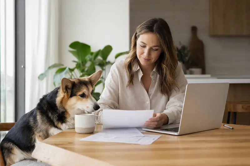 Ein ruhiger Corgi sitzt neben seinem Besitzer am Küchentisch, auf dem Versicherungsunterlagen und ein Laptop liegen.