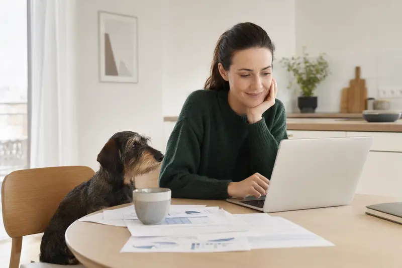 Ein ruhiger Dachshund sitzt neben seinem Besitzer am Küchentisch, während Versicherungsunterlagen und ein Laptop auf dem Tisch liegen.