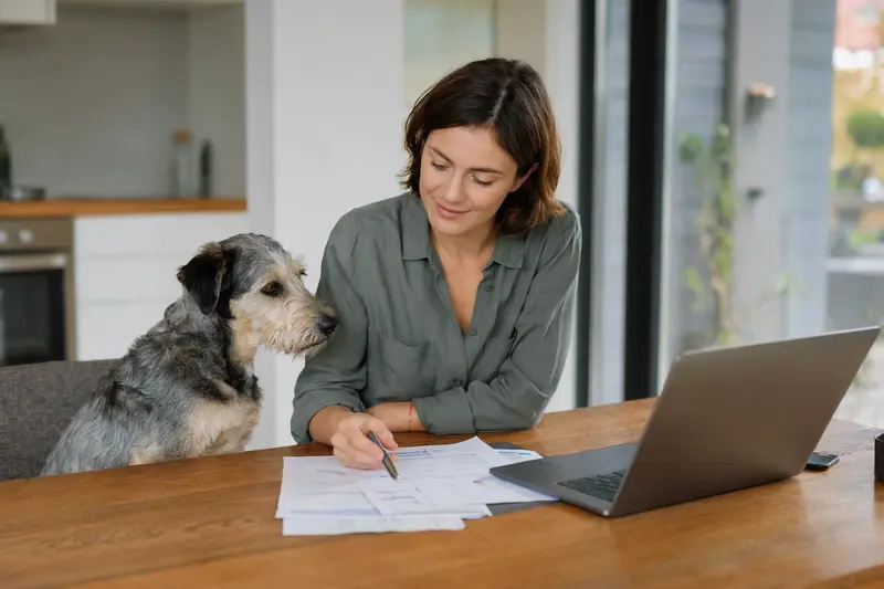Ein Designerhund sitzt entspannt neben seinem Besitzer am Küchentisch, auf dem Versicherungsunterlagen und ein Laptop liegen.