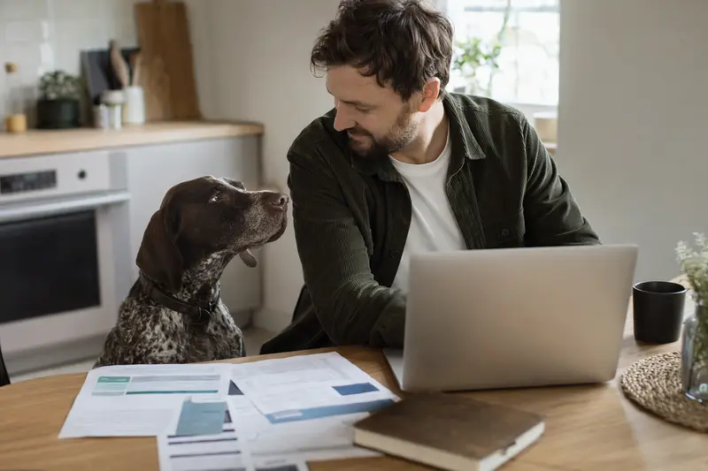 Ein Deutsch-Kurzhaar Hund sitzt neben seinem Besitzer am Küchentisch, während auf dem Tisch Versicherungsunterlagen und ein Laptop liegen.