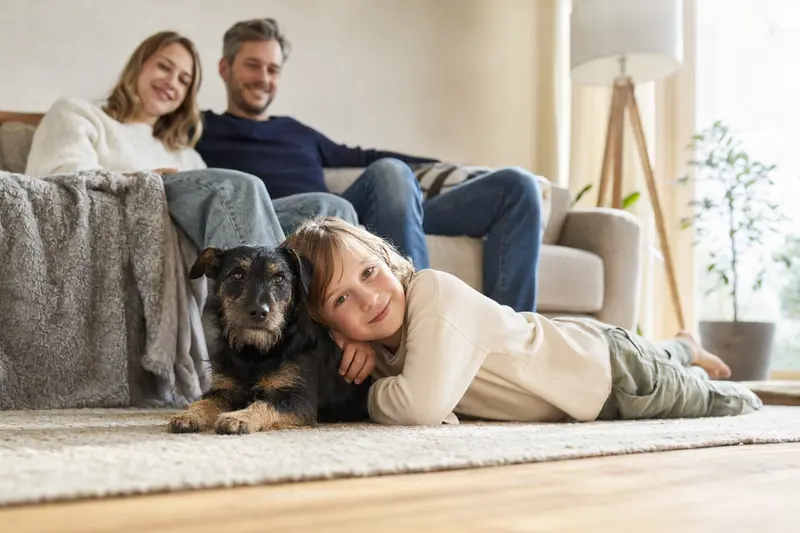 Ein Deutscher Jagdterrier liegt entspannt neben einem Kind im Wohnzimmer, während die Eltern im Hintergrund eine sichere Familienatmosphäre schaffen.