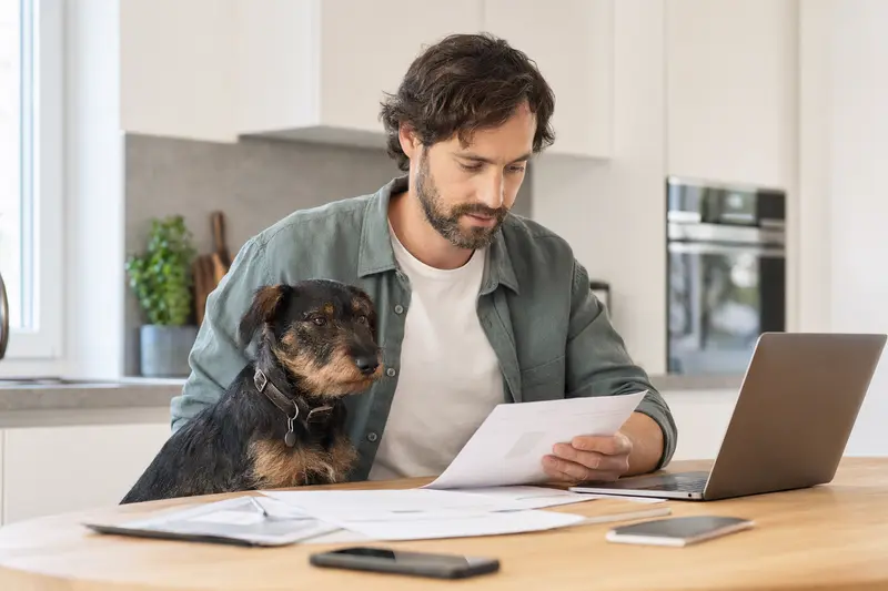 Ein Deutscher Jagdterrier sitzt ruhig neben seinem Besitzer am Küchentisch mit Versicherungsunterlagen und einem Laptop zur Hundeversicherung.