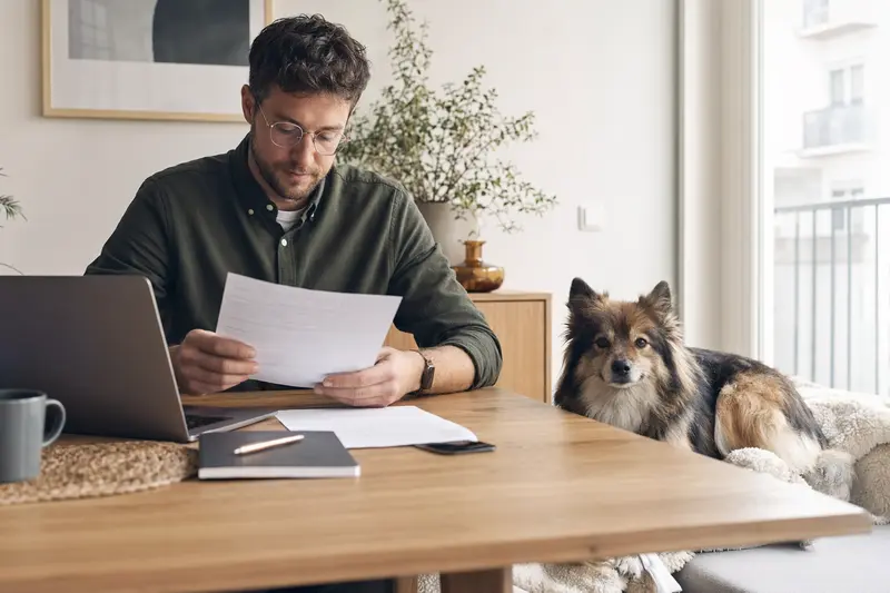 Ein Hundebesitzer sitzt mit einer Tierarztrechnung am Tisch, während ein Deutscher Spitz entspannt auf einer Decke liegt – ein ruhiges Motiv zu Tierarztkosten und Versicherung.