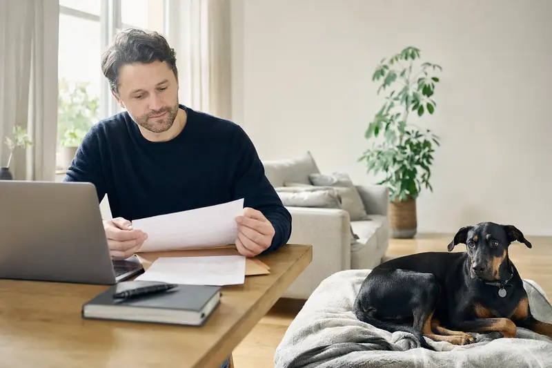 Ein Hundehalter sitzt mit einer Tierarztrechnung am Tisch, während ein entspannter Dobermann auf einer Decke neben ihm liegt.