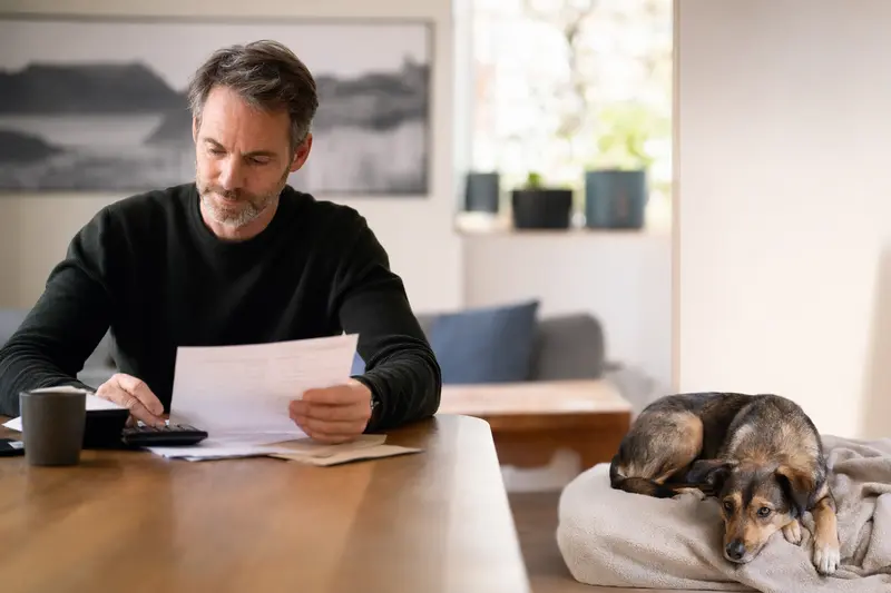 Ein Hundebesitzer sitzt mit einer Tierarztrechnung am Tisch, während ein entspannter Doguillo auf einer Decke liegt und den ruhigen Umgang mit Tierarztkosten im Versicherungsalltag zeigt.