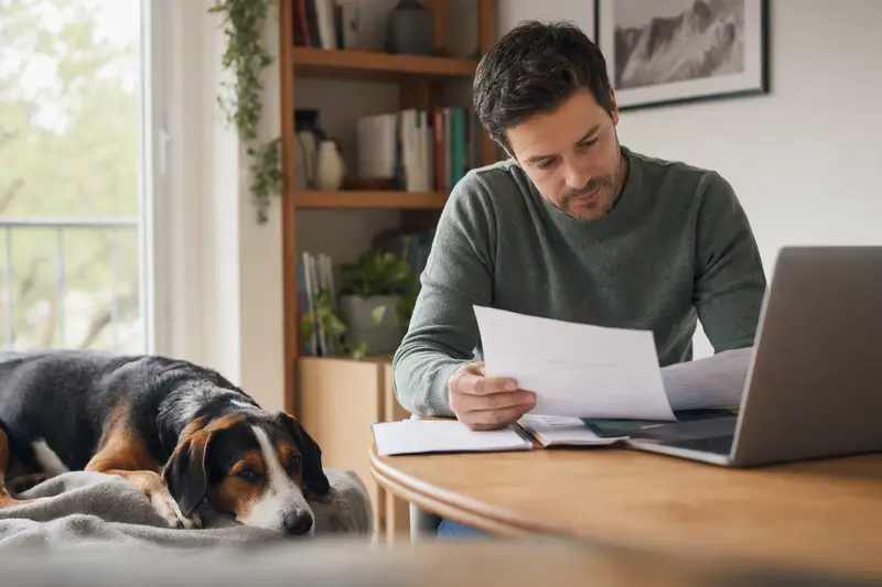 Ein Hundebesitzer sitzt mit einer Tierarztrechnung am Tisch, während ein dreifarbiger Serbischer Laufhund entspannt auf einer Decke liegt.