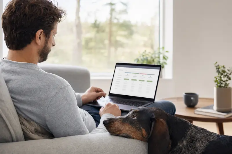 Ein Hundehalter sitzt mit seinem Dunkerbracke Hund auf dem Sofa und vergleicht Versicherungsoptionen auf dem Laptop in einer ruhigen, hellen Wohnung.