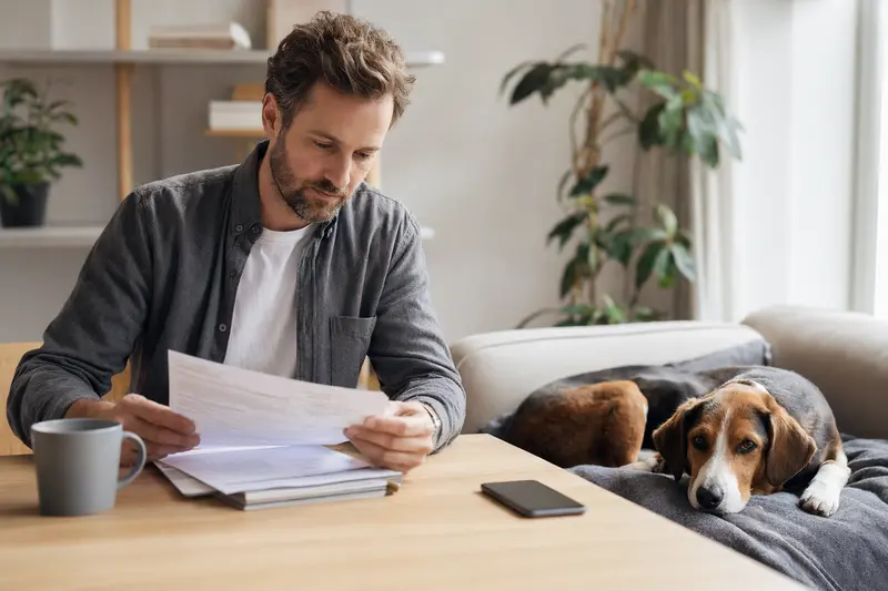 Ein Hundehalter sitzt mit Tierarztrechnung am Tisch, während ein entspannter Finsk Stövare auf einer Decke liegt und das Thema Hundekrankenversicherung ruhig und sachlich vermittelt.