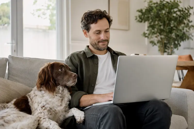 Ein Hundebesitzer sitzt mit einem Laptop auf dem Sofa, während ein Französischer Spaniel daneben liegt und eine neutrale Versicherungsvergleichs-Oberfläche sichtbar ist.