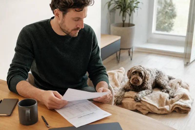 Ein Hundebesitzer sitzt mit einer Tierarztrechnung am Tisch, während ein entspannter Französischer Wasserhund ruhig auf einer Decke liegt und das Thema Tierarztkosten und Absicherung im Alltag zeigt.