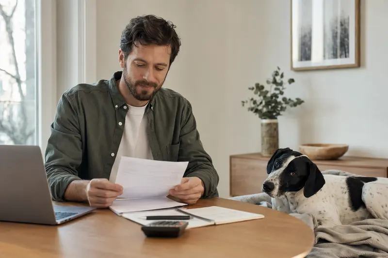 Ein Hundebesitzer sitzt mit einer Tierarztrechnung am Tisch, während ein entspannter Französischer Weiß-Schwarzer Laufhund auf einer Decke daneben ruht.