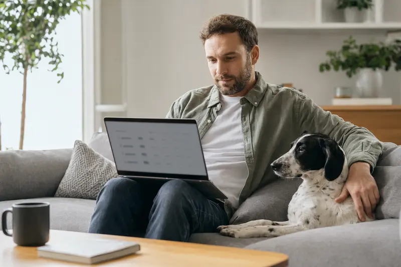 Ein Hundebesitzer arbeitet am Laptop auf dem Sofa, während ein Französischer Laufhund entspannt daneben sitzt.