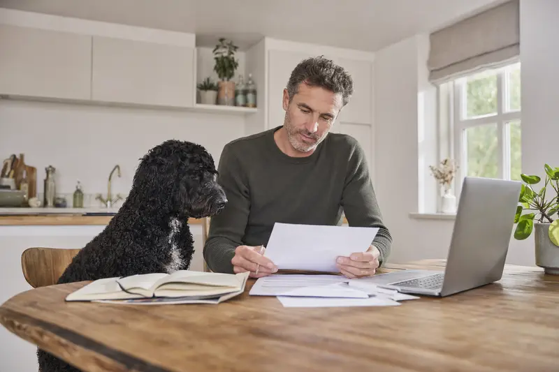 Friesischer Wasserhund sitzt neben seinem Besitzer am Küchentisch mit Versicherungsunterlagen und einem Laptop.